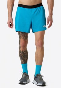 Turquoise athletic shorts with a black waistband, featuring side slits. A prominent tattoo on one leg. Paired with gray shoes and blue socks.