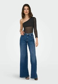 Sort asymmetrisk mesh bodysuit parret med højtaljede, vide blå denimbukser. Blød tekstur og karakteristisk transparent ærmedetail.