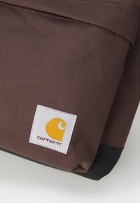 Brown fabric backpack with a textured surface and a black bottom, featuring a white label with a yellow logo. Simple, functional design.