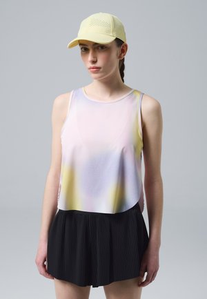 Giovane donna con top smanicato color pastello, gonna nera plissettata e cappellino giallo chiaro, in piedi davanti a uno sfondo grigio uniforme.