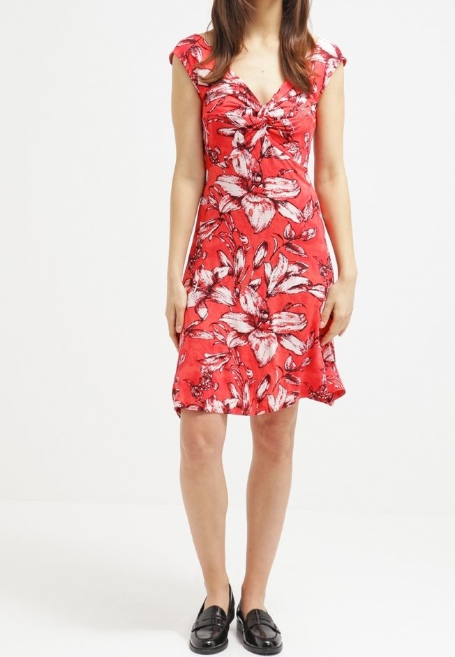 Femme portant une robe fleurie rouge mi-longue aux manches courtes et des mocassins noirs, se tenant devant un fond blanc uni.