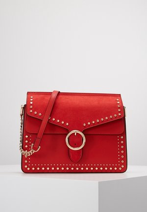 Rode leren handtas met gouden studs die de klep en randen omlijnen, gouden ronde gesp en een kettingriemdetail op een witte ondergrond.