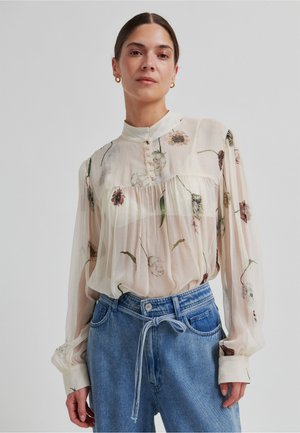 Przezroczysta kremowa bluzka w kwiatowe wzory, z wysokim kołnierzem i patką na guziki. Długie rękawy z kontrastowymi mankietami w połączeniu z dżinsami.
