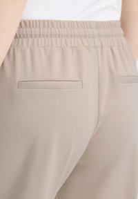 Pantalon beige à taille élastique avec deux poches horizontales à l'arrière, porté avec un haut blanc, vu de la taille aux hanches.