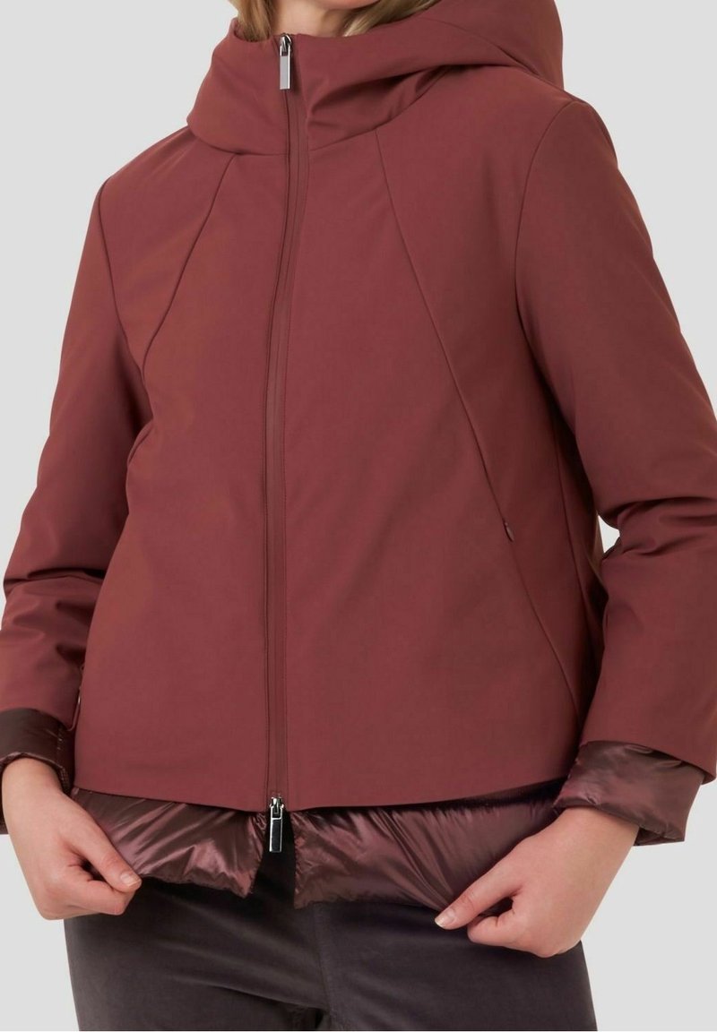 Personne portant une veste à capuche bordeaux avec une fermeture éclair à l'avant et une doublure intérieure matelassée, ajustant l'ourlet de la veste avec les deux mains.
