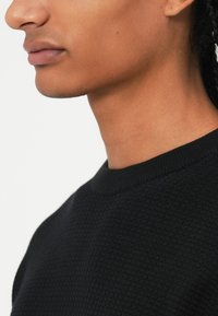 Maglione nero a maglia con scollo rotondo, caratterizzato da un motivo testurizzato e dettagli a costine sul colletto. Vista ravvicinata del tessuto.