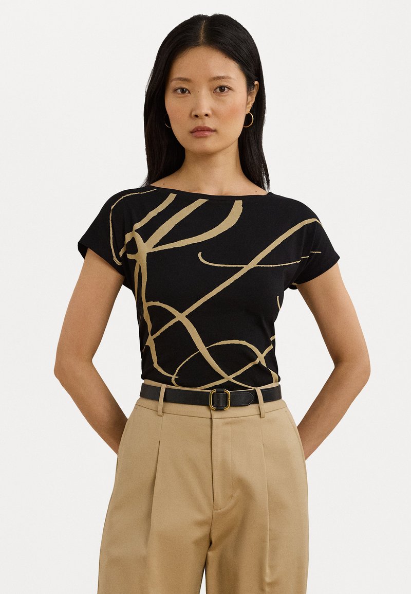 Camiseta negra con diseño abstracto en línea dorada, de mangas cortas y cuello redondo, combinada con pantalones beige de tiro alto y un cinturón negro.