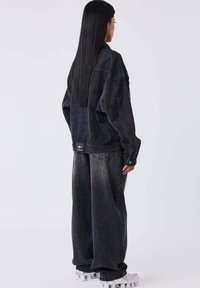 Giacca in denim scuro oversize, pantaloni abbinati larghi, entrambi con effetti di scolorimento. Accenti in metallo argentato e un patch con logo sul retro.