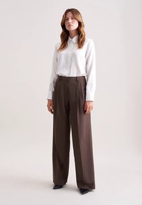 Biała koszula w paski, zapinana na guziki, wpuszczona w brązowe, szerokie spodnie z wysokim stanem. Strój ma gładką fakturę i dopasowany krój.