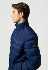 Navy puffjakke har en høj krave, vandret quiltet design og et brun læderlogo på venstre ærme. Glat, skinnende tekstur.
