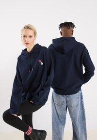 Μπλε ναυτικού φούτερ με κουκούλα σε παχύ ύφασμα, που διαθέτει εκτυπωμένο σχέδιο με ένα χέρι που κρατάει μια καρδιά. Χαλαρή εφαρμογή, καγκουρό τσέπη και κουκούλα με κορδόνι.