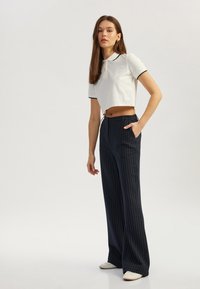 Witte cropped poloshirt met zwarte randen, gecombineerd met een marineblauwe pinstriped broek met wijde pijpen. Model poseert met handen in de zakken.