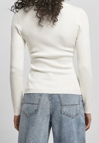 Prążkowany, długi rękaw w kolorze kremowym, wykonany z miękkiego, elastycznego materiału. Połączony z jasnoniebieskimi, wysokim stanem jeansami z tylnymi kieszeniami.