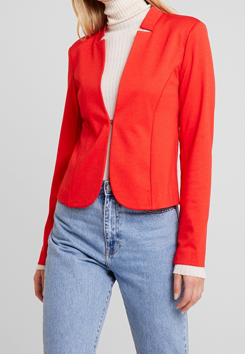 Blazer rouge ajusté avec une fermeture à deux boutons, texture lisse et manches longues, associé à un col roulé crème et un jean bleu clair.
