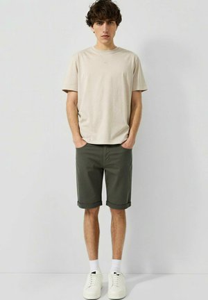 Jeune homme debout, mains dans les poches, portant un t-shirt beige, un short vert foncé retroussé, des chaussettes blanches et des baskets blanches devant un mur uni.