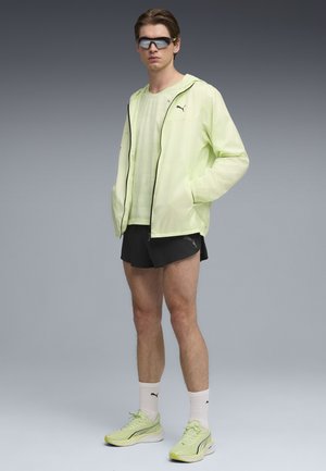 Hombre con chaqueta y zapatos Puma de color amarillo claro, pantalones cortos negros, calcetines blancos Puma y gafas de sol oscuras, de pie frente a un fondo gris.