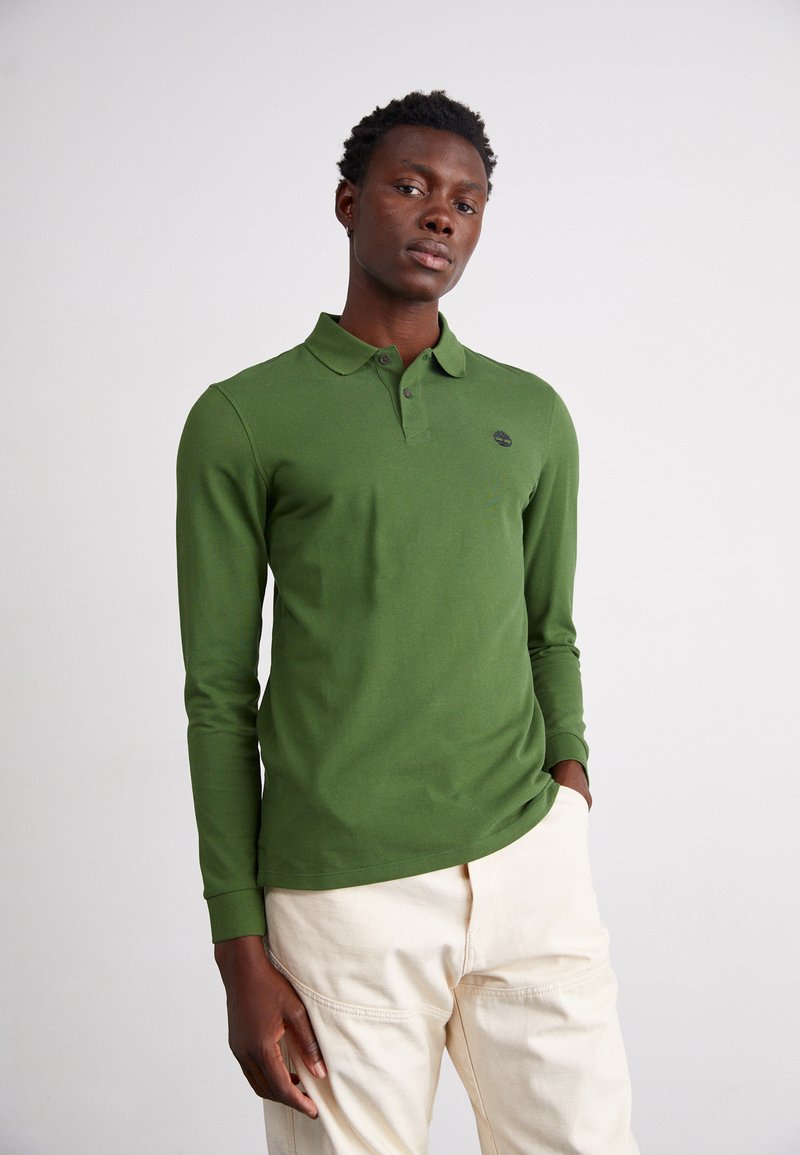 Camisa polo verde de manga longa com colarinho clássico, pala de botão duplo e detalhe de logotipo; combinada com calças beje claras. Textura suave.