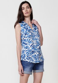 Blusa senza maniche con un motivo a foglie blu e bianche, colletto, chiusura con bottoni e tessuto leggero, abbinata a pantaloni corti di jeans arrotolati.
