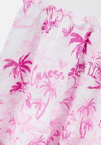 Vaaleanpunainen ja valkoinen kukkamekko, jossa on palmupuukuvioita, kevyt kangas, röyhelöinen pääntie ja vaaleanpunainen "Guess"-logopainatus.