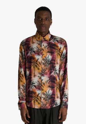 Uomo che indossa una camicia a maniche lunghe con bottoni e motivo di palme arancioni, rosa, bianche e nere, in piedi davanti a uno sfondo bianco.