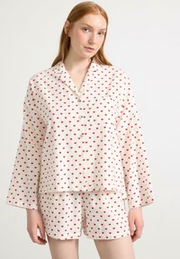 Vitt pyjamaset med röda hjärtan. Löst passande, långärmad topp med knappstängning och matchande shorts. Mjuk, lättviktsstoff.