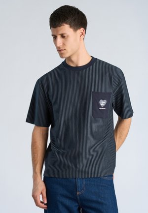 Junger Mann trägt ein dunkelblaues T-Shirt mit Nadelstreifen und einer dunklen Tasche mit herzförmigem Design, kombiniert mit blauen Jeans, schaut nach unten.