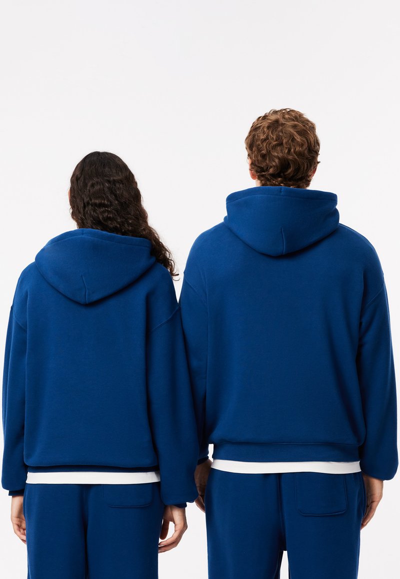 Sudaderas con capucha azul de corte holgado, con puños acanalados y un bolsillo frontal. Ambas están combinadas con pantalones azules a juego.