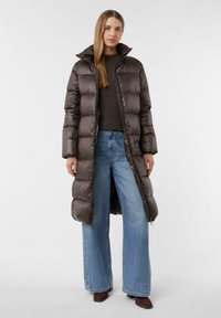 Brauner Daunenmantel mit hohem Kragen, glänzender gesteppter Textur, langem Schnitt und Reißverschluss. Kombiniert mit blauen weiten Jeans.