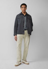 Donkergrijze jas met twee borstzakken, gedragen over een grijze trui. Off-white broek en lichte sneakers met een zandkleurige zool en zwarte accenten.