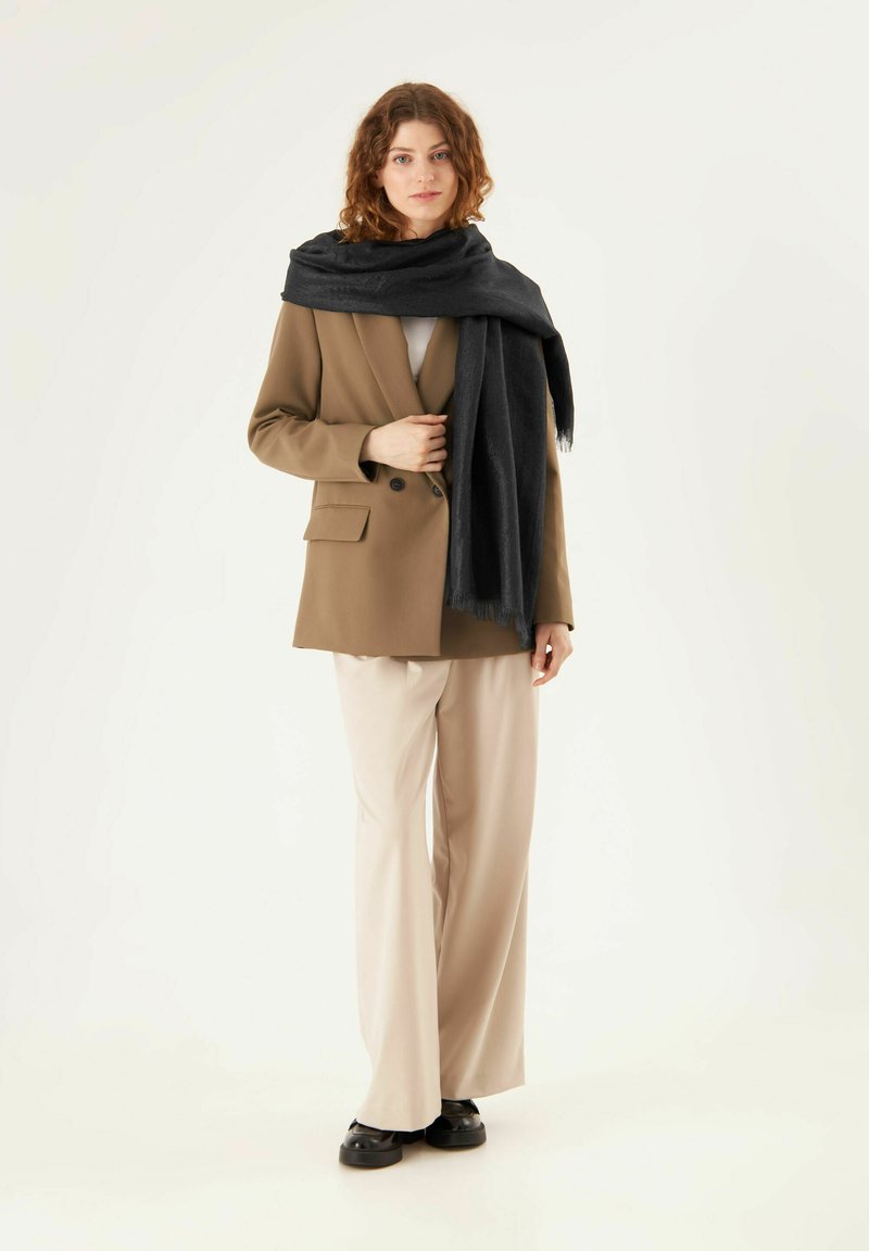 Bruine op maat gemaakte blazer over een witte top, gecombineerd met beige wijde pantalon en een zwarte sjaal. De outfit kenmerkt zich door een minimalistisch ontwerp en texturen.