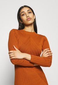 Narančasta haljina s rebrastim uzorkom, dugim rukavima i visokim izrezom, ukrašena geometrijskim uzorcima. Ima teksturiranu površinu i uski kroj.