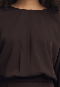 Pruun särk, millel on sile tekstuur, laevanina ning voltimise detailid. Kangas jäseb lõdvalt torso ümber.