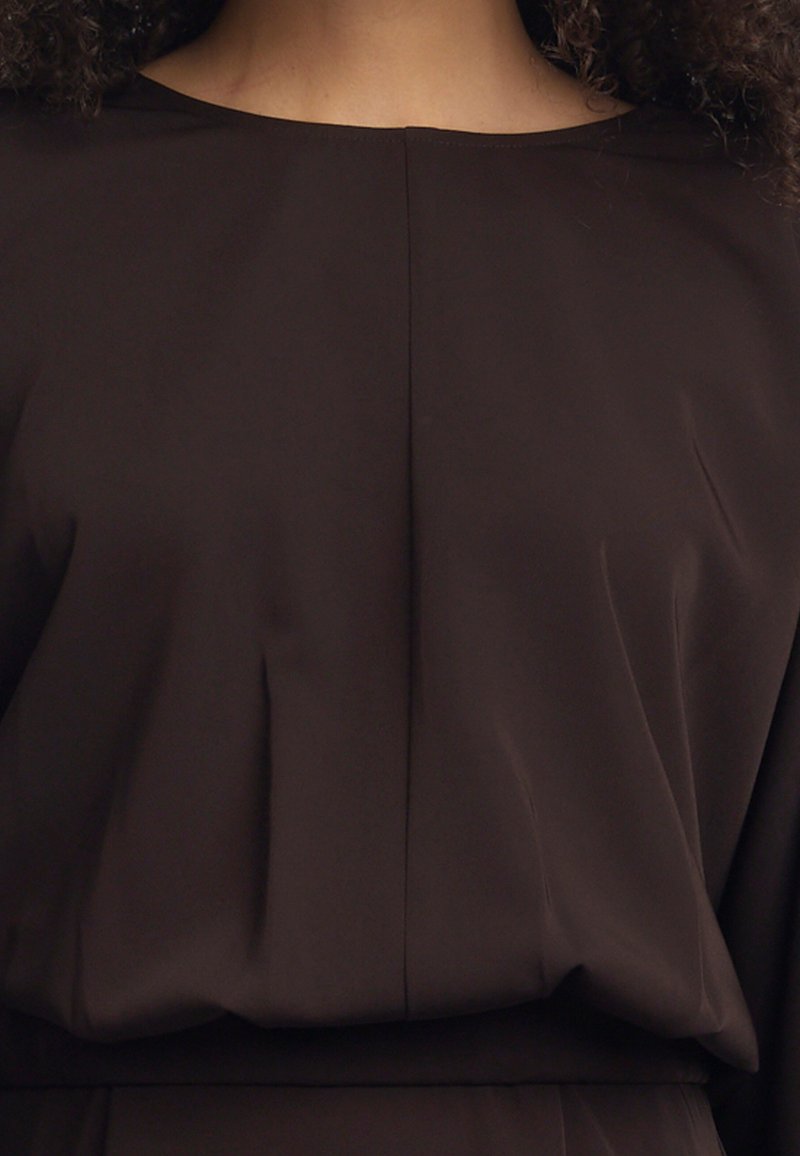Braune Bluse mit glatter Textur, ausgestattet mit einem Bootsausschnitt und Fälteldetails. Der Stoff fällt locker um den Oberkörper.
