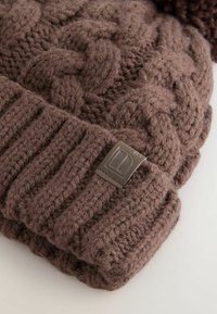 Brown knitted beanie with a braided pattern, featuring a ribbed cuff and a leather logo patch. Soft, textured wool material.