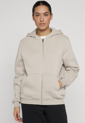 Kvinde iført beige hættetrøje med lynlås og lommer, hænderne i lommerne, foran en ensfarvet, lyse baggrund.