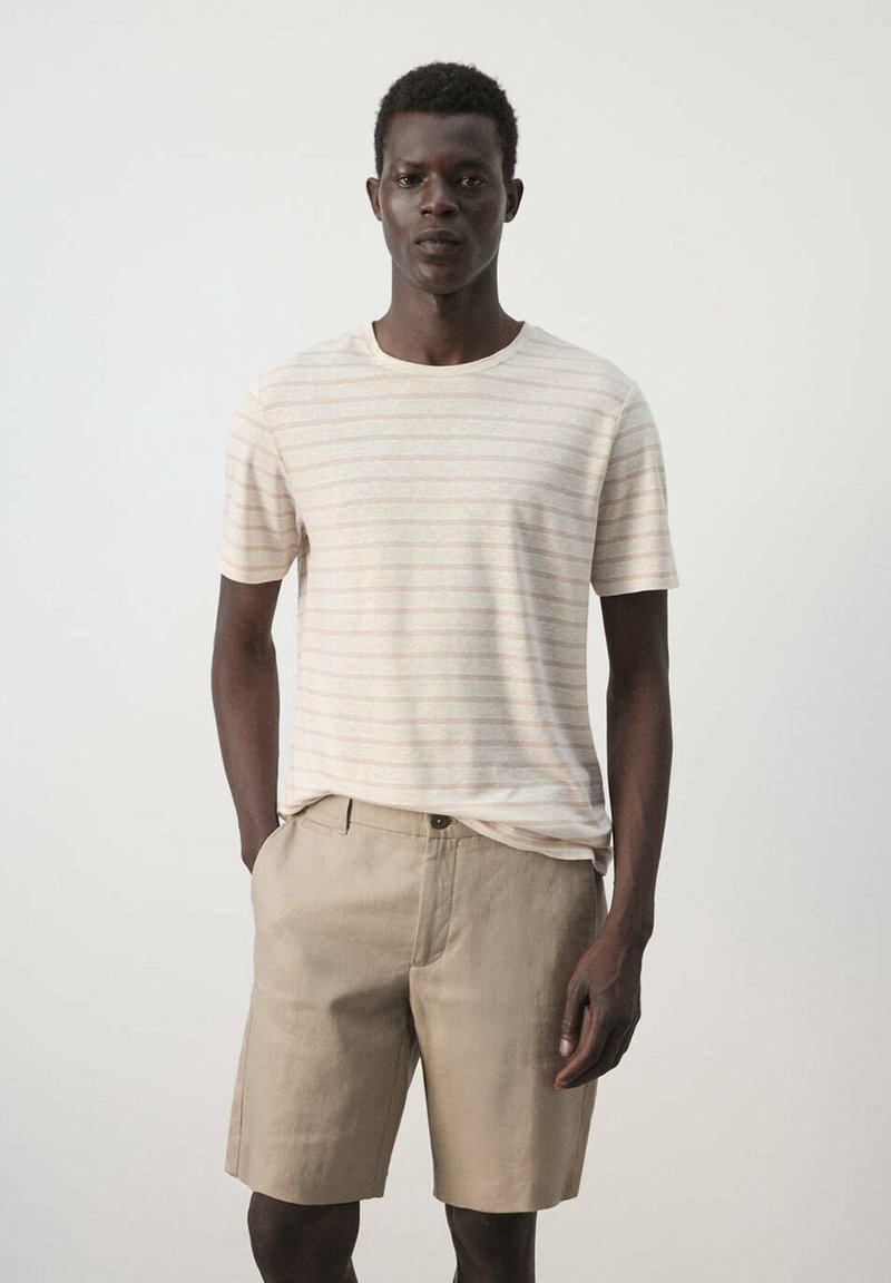 Homme debout les mains dans les poches portant un t-shirt rayé beige et un short marron clair sur un fond uni.