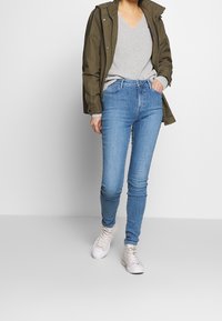 Jean en denim bleu clair à coupe slim, accompagné d'un pull en maille gris et d'une parka vert foncé. Des baskets blanches complètent la tenue.
