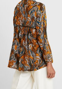 Blusa a maniche lunghe, arancione e a fantasia con design paisley, vista da dietro, abbinata a pantaloni bianchi.