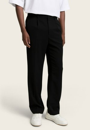 Osoba ubrana w czarne, dopasowane spodnie, białe trampki i białą koszulę z krótkim rękawem, stojąca na tle jednolitego tła.