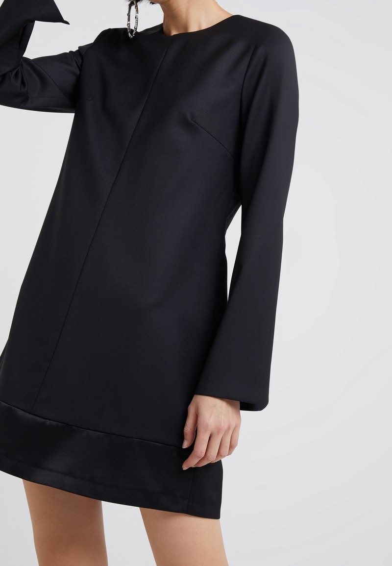 Femme portant une mini-robe noire à manches longues avec un col rond et de larges poignets, debout devant un fond clair uni.