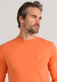 Oranger Strickpullover mit geripptem Kragen und langen Ärmeln. Verfügt über ein kleines Logo in einem helleren Farbton auf der Brust. Weicher Stoff.