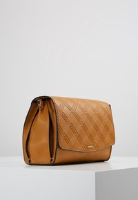 Borsa a mano in similpelle color tan con motivo trapuntato, chiusura a patta e piping nero. Presenta una tasca con zip sul lato per ulteriore spazio di stoccaggio.