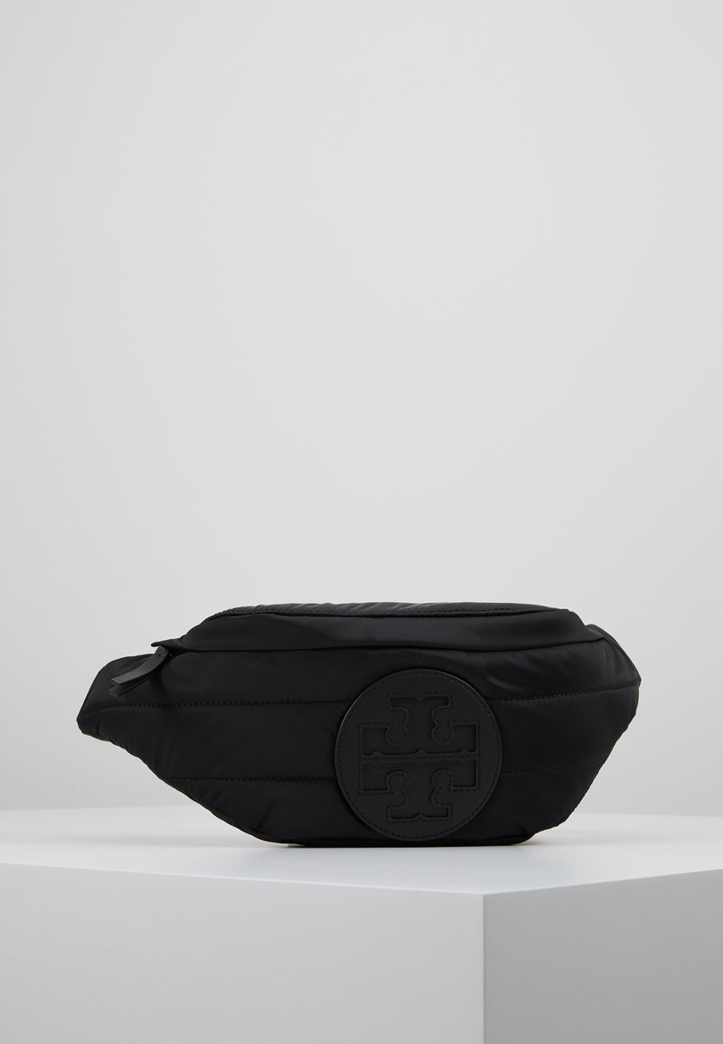Sac banane noir matelassé avec fermeture éclair à l'avant et logo rond brodé, placé sur une surface d'exposition blanche sur fond gris uni.