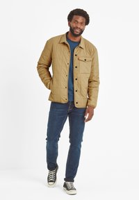 Tan quilted jacket with a button-up front, chest pocket, and black accents, worn over a blue shirt, paired with dark jeans and sneakers.