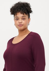 Woman with curly hair in a high bun wearing silver hoop earrings and a long-sleeve maroon top, facing forward against a plain white background.