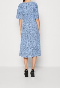 Abito midi blu con stampa floreale, maniche corte, vita arricciata e silhouette morbida. Indossato con scarpe col tacco nere.