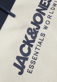 Tela beige con texto en azul marino que dice "JACK & JONES ESSENTIALS WORLDWIDE." Presenta un acabado texturizado y extremos de cordón redondeados.