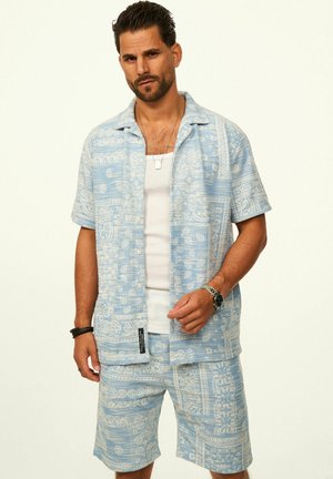 Man wearing light blue patterned short-sleeve shirt and matching shorts over white tank top, with wristwatch and necklace, standing against white background.