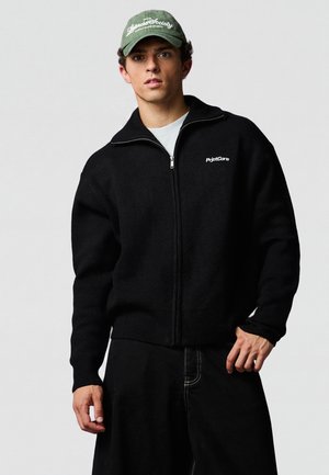 Junger Mann mit grüner Baseballkappe und schwarzem Zip-Up-Pullover mit weißem Logo, steht vor einem einfarbigen hellgrauen Hintergrund.
