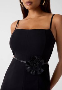 Schwarzes Kleid mit quadratischem Ausschnitt, dünnen Trägern und tailliertem Oberteil. Mit einer Lederblumen-Applikation in der Taille und einer glatten Textur.